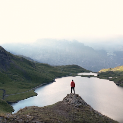 Eine Person in roter Jacke steht auf einer felsigen Bergspitze über einem ruhigen alpinen See, umgeben von grünen Hügeln und hohen, teilweise im Nebel liegenden Bergen.